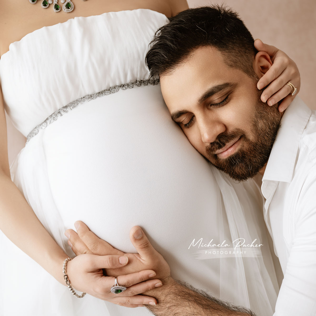 Mann lehnt mit geschlossenen Augen liebevoll an den Babybauch seiner schwangeren Partnerin, beide halten ihre Hände über dem Bauch.